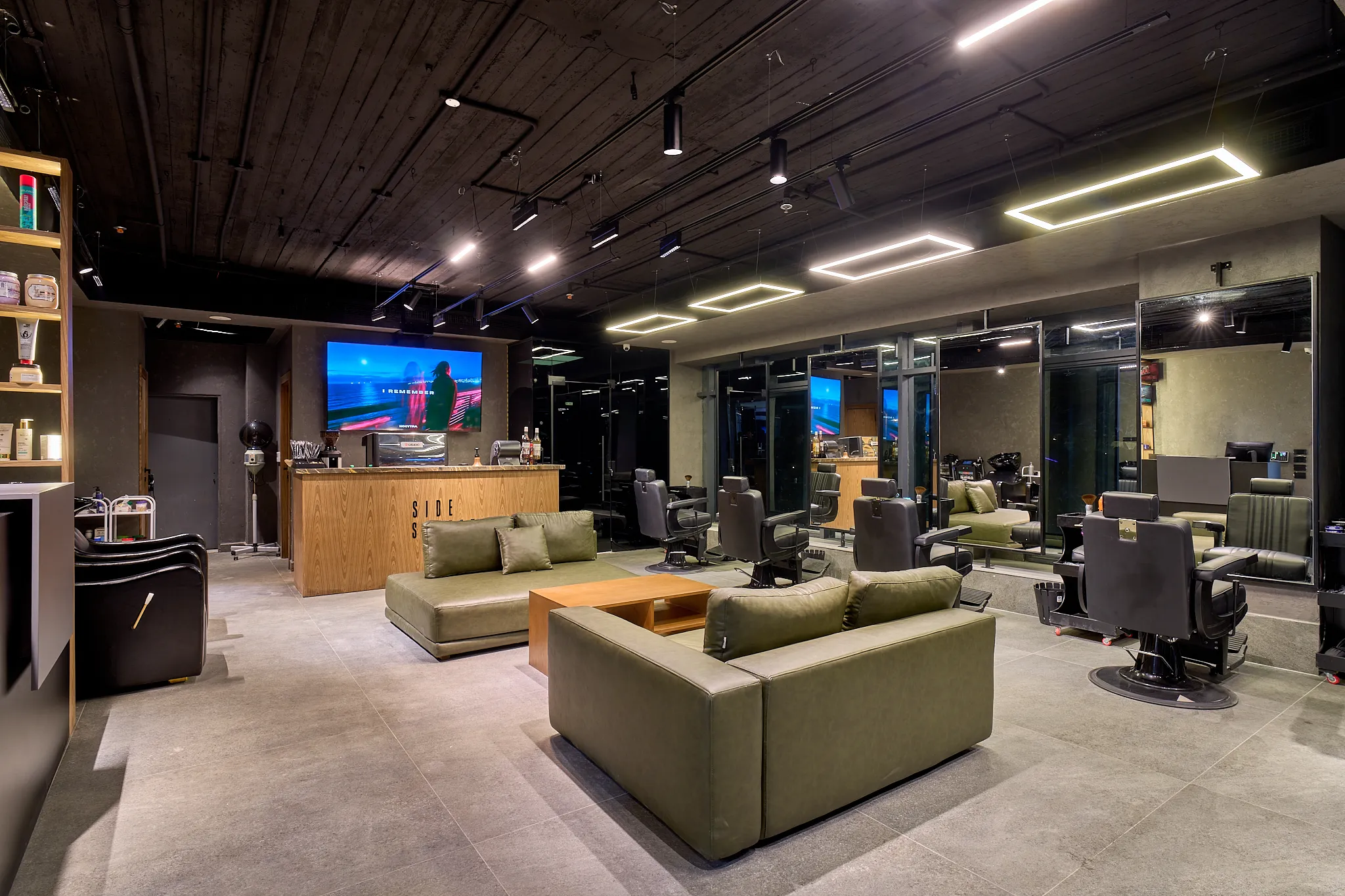 The main hall of KK Barbershop: olive-green leather sofas around a wooden coffee table, a wood-clad coffee counter beneath a wall screen, and black barber chairs facing mirrors on the right, under a dark timber ceiling with linear lights.