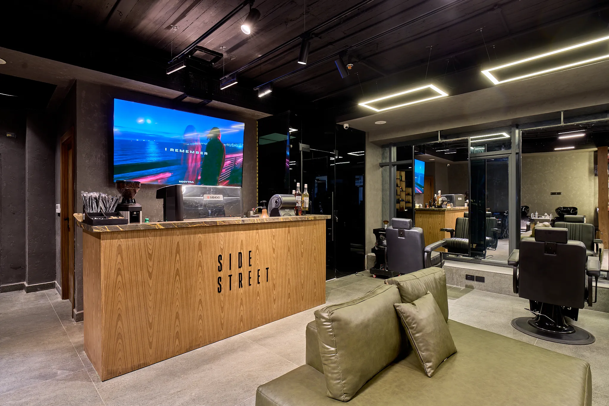 The wood-clad coffee counter with an espresso machine and a grinder below a wall screen, an olive-green sofa in the foreground and barber chairs to the right.