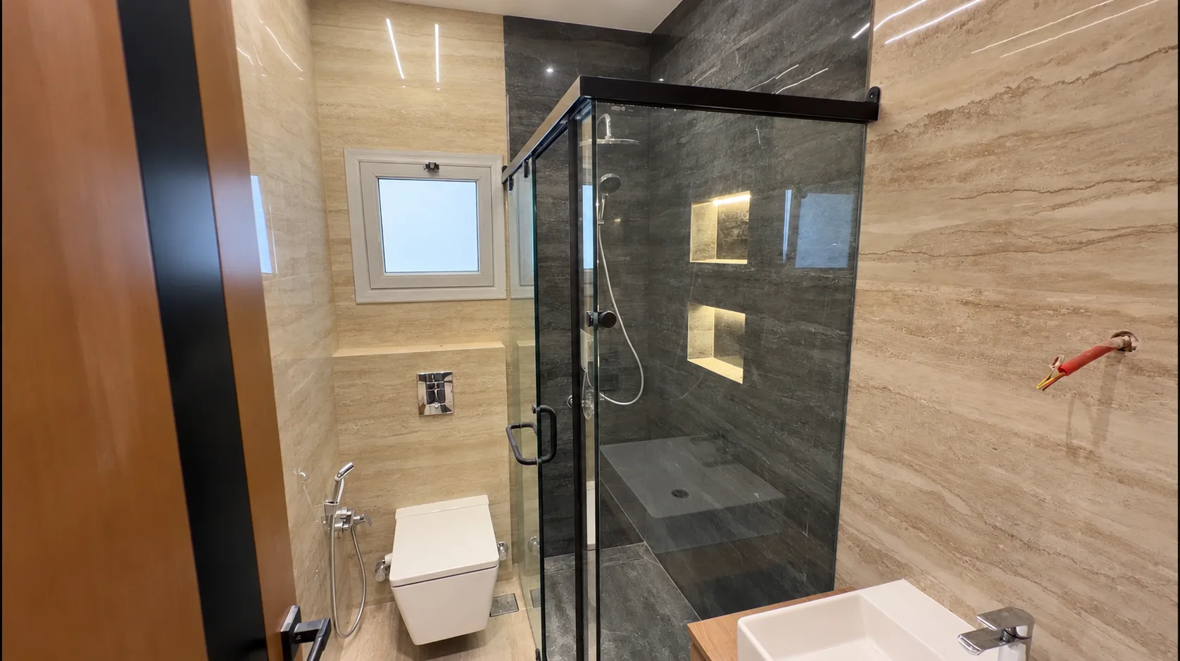 A bathroom with travertine-look walls, a glass shower with lit niches and a wall-hung toilet.