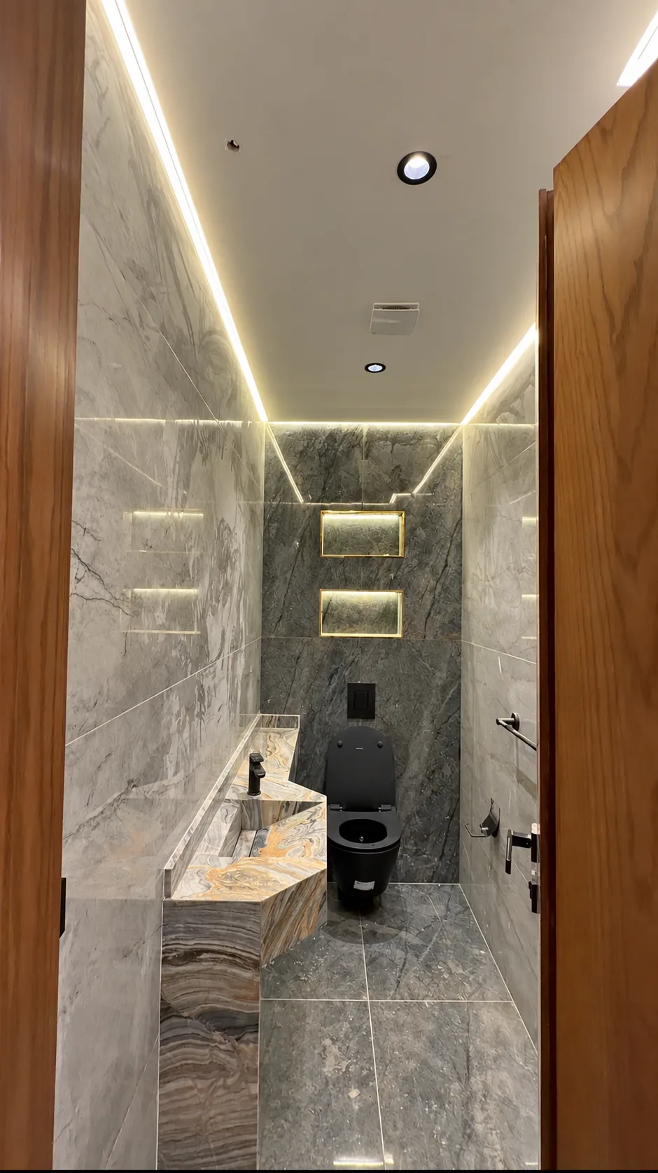 A washroom in grey veined stone with a black toilet, a stone basin and a lit cove.